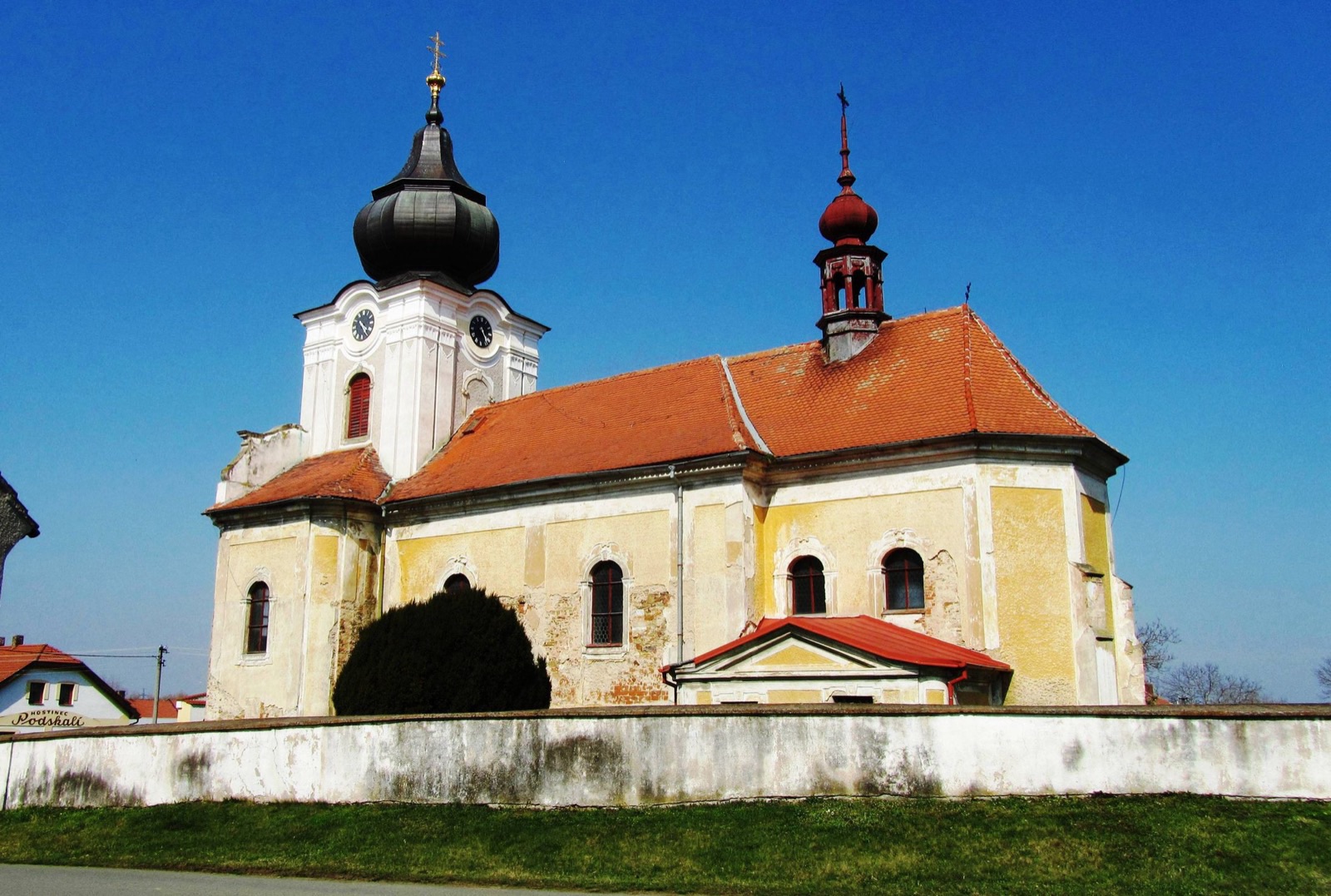 Žlutý barokní kostel sv. Vavřince s cibulovou věží za bílou ohradní zdí